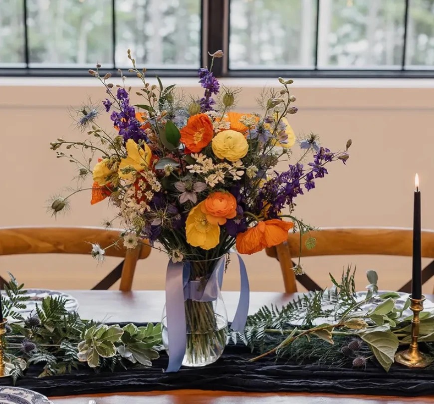 A wildflower centerpiece of poppies, ranunculus, and delphinium in a glass vase on the reception table