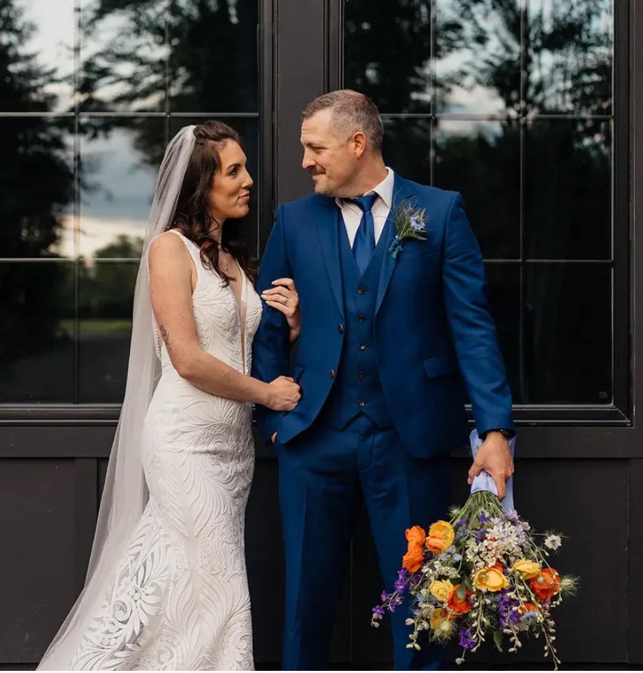 The bride and groom sharing a quiet look against the venue's black windowed doors, a wild bouquet of poppies and delphinium in hand