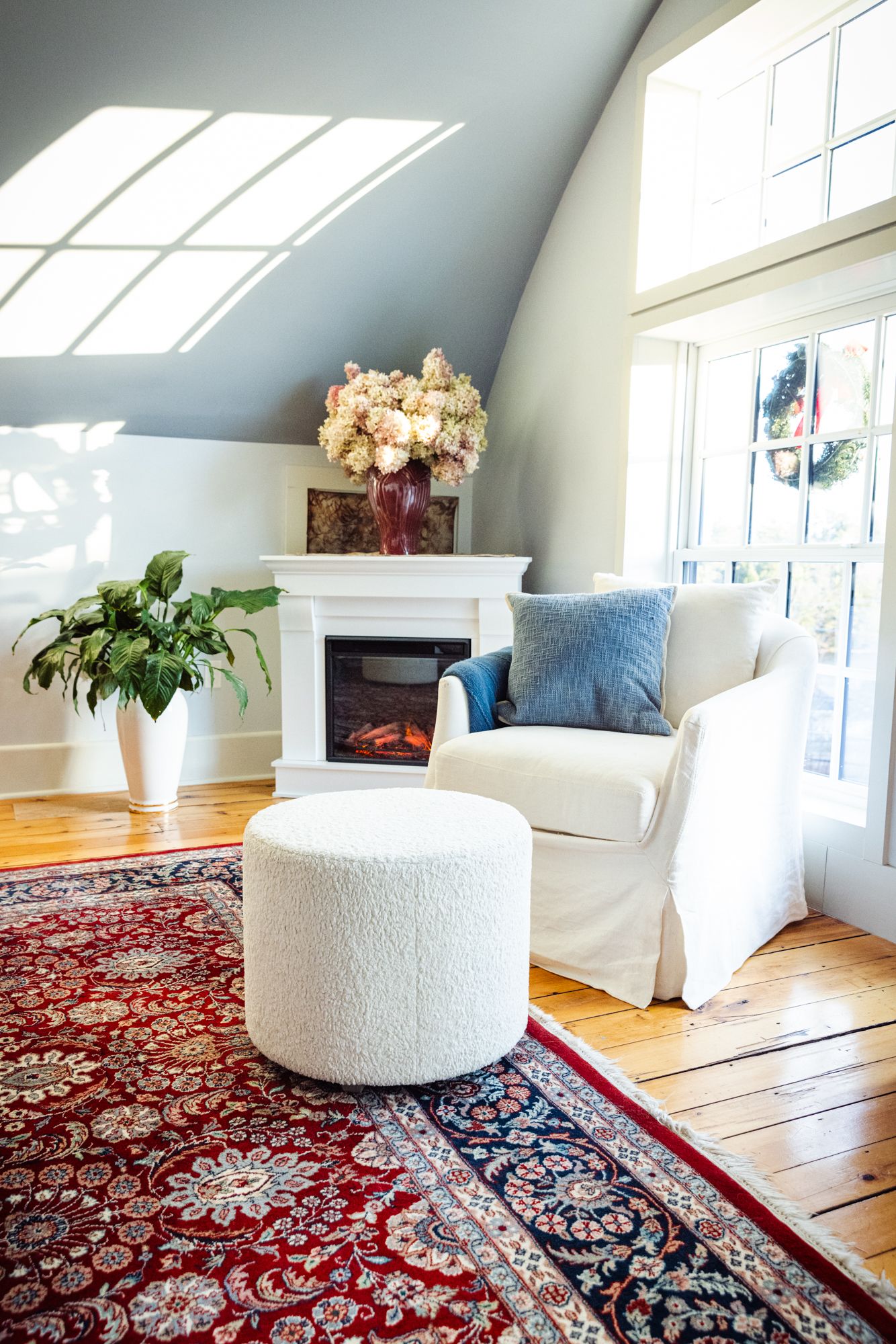 The primary suite's sitting corner — a fireplace, dried hydrangeas, and a linen chair beneath the vaulted ceiling